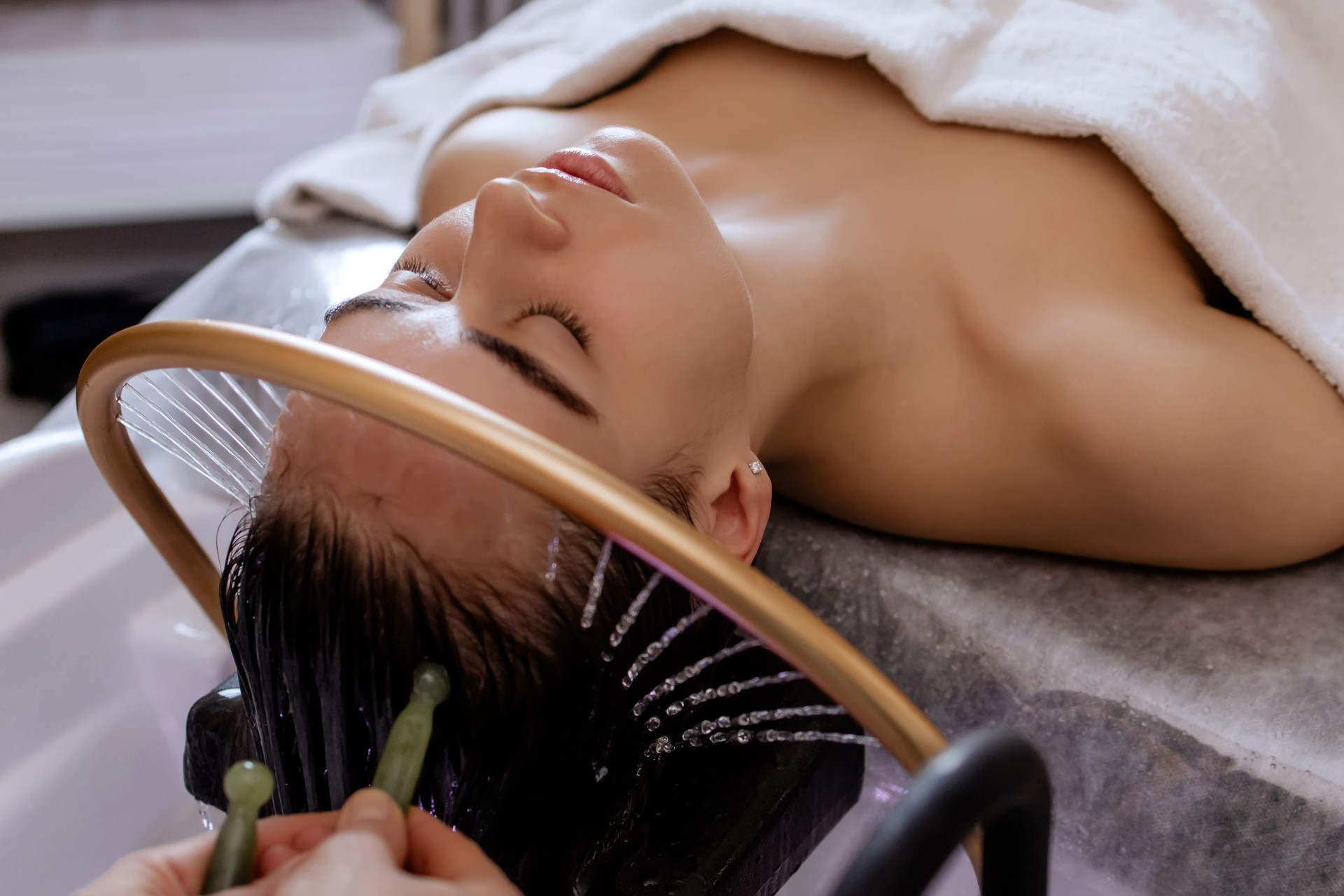 Close-up of a woman receiving a relaxing Japanese head spa treatment, with green massage sticks applied to her scalp, while water gently flows from a special tool during the session in a beauty salon